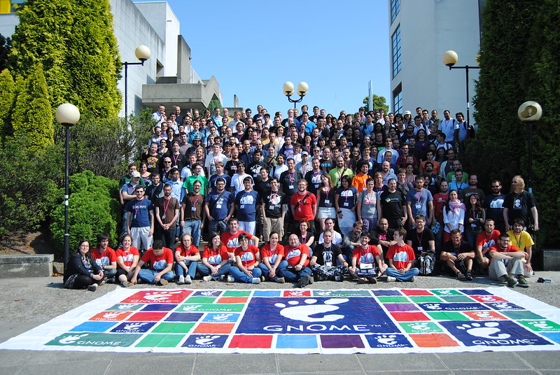 This photo includes a large group of people standing on steps outside on a sunny day. In the foreground is a large beach blanket with brightly colored GNOME logos on it.